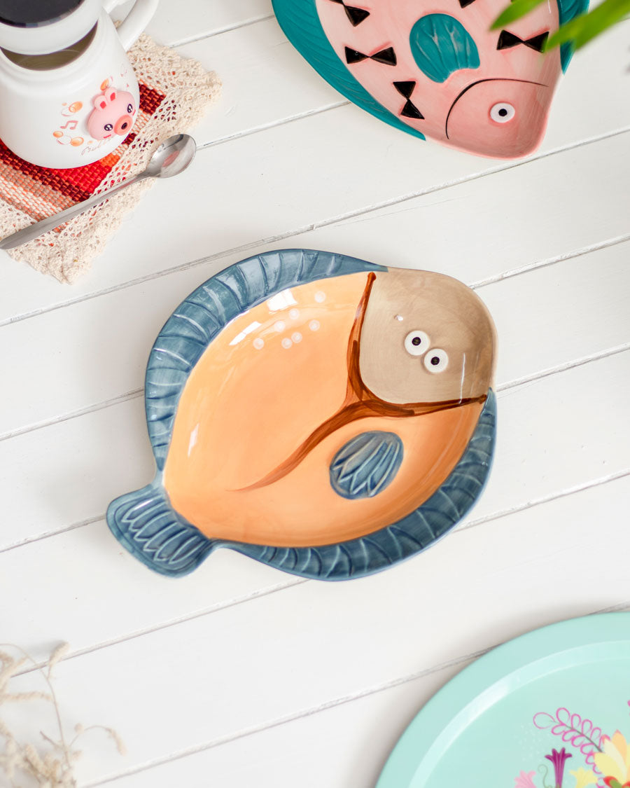 Orange 'Gilligan' fish-shaped ceramic platter alongside a cup and saucer on a white wooden surface.