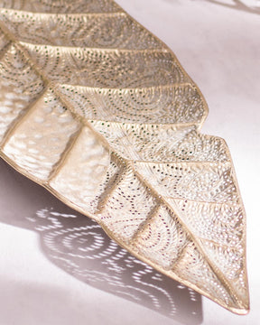 An intricately designed metal leaf serving platter with a textured, gold-finished surface. The platter is shaped like an elongated leaf with detailed vein patterns and lace-like cutouts, casting a delicate shadow on a light surface.