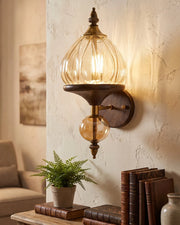A wall-mounted sconce with a ribbed clear glass dome shade, a warm glowing bulb, and a dark bronze metal base mounted on a textured beige wall in a styled interior setting.